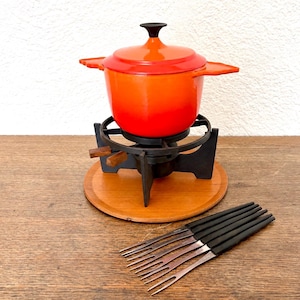 May include: A bright orange enameled cast iron fondue pot with a matching lid and black knob, resting on a black metal stand with wooden handles, set upon a round wooden base, accompanied by six metal fondue forks with black plastic handles.