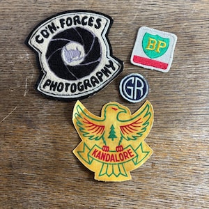 May include: A collection of embroidered patches with various designs. One patch features a camera lens and the text "CON. FORCES PHOTOGRAPHY." Another has a green shield with "BP." A yellow patch displays an eagle and the text "KANDALORE."