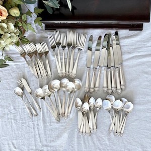 Puede incluir: Una colección de cubiertos plateados antiguos, que incluye tenedores, cuchillos, cucharas y cucharas de servir. La cubertería está dispuesta sobre un mantel gris claro. Los mangos tienen un diseño floral decorativo.