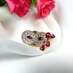 Puede incluir: Un broche de tono dorado con una máscara de pedrería blanca con detalles de pedrería roja y un lazo rojo.