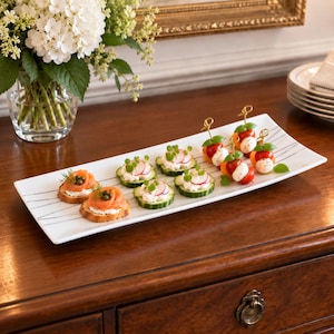 Może przedstawiać: Prostokątna taca do serwowania z białej ceramiki z cienkimi, ciemnoszarymi ukośnymi liniami. Na tacy znajduje się asortyment przekąsek, w tym wędzony łosoś na chlebie, plasterki ogórka z serkiem śmietankowym i szaszłyki caprese.