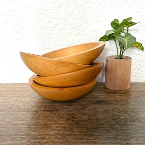 May include: Four wooden bowls stacked on top of each other. The bowls are a light brown colour and have a smooth, rounded shape. The bowls are sitting on a wooden table.