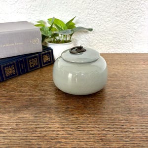 以下が含まれることがあります： 蓋と濃い金属製のリングハンドルが付いた、小さくて丸い、明るい灰色のセラミックジャー。ジャーは、本と小さな鉢植えの植物の隣の木製の表面に置かれています。