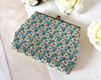 May include: A vintage beaded clutch bag with a floral design. The bag has a light grey base with a repeating pattern of blue and green flowers. The frame is bronze-coloured, and the clasp is closed. White roses are in the background.