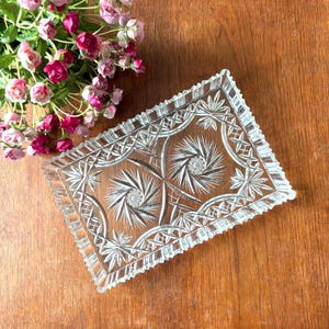 Puede incluir: Una bandeja rectangular de vidrio transparente con intrincados diseños de estrellas y geométricos. La bandeja tiene un borde festoneado y probablemente sea vintage. Está sobre una superficie de madera, con flores rosas y moradas en el fondo.