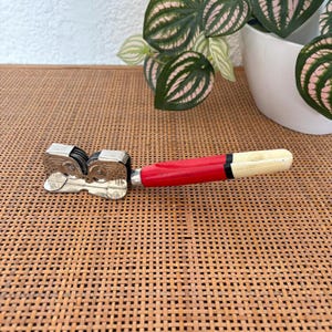 May include: A manual knife sharpener featuring a metallic head with two circular grinding wheels and a handle composed of a bright red section followed by a shorter, cream-coloured end piece, separated by a thin black band.