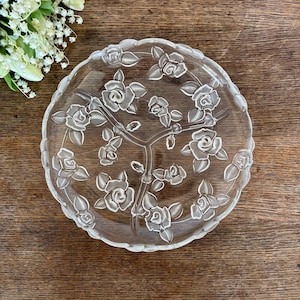 Puede incluir: Plato de servir de vidrio transparente con tres compartimentos, con un diseño de rosas en relieve. El plato tiene un borde festoneado y está sobre una superficie de madera. Ideal para servir aperitivos o postres.