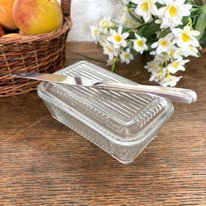 May include: A clear glass butter dish with a ribbed lid and a silver butter knife. The dish is rectangular and sits on a wooden surface. A basket of fruit and flowers are in the background.