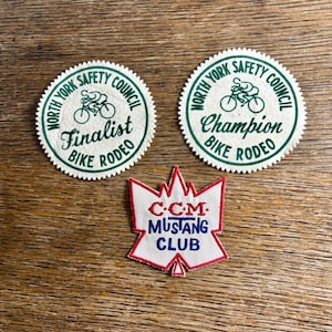 Pode incluir: Três patches vintage em uma superfície de madeira. Dois patches circulares com o texto "North York Safety Council" e "Bike Rodeo", um com a leitura "Finalist" e o outro "Champion". Um patch vermelho, branco e azul do "CCM Mustang Club" também está presente.