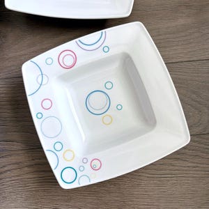 Puede incluir: Cuenco cuadrado de cerámica blanca con esquinas redondeadas. El cuenco está decorado con círculos de colores azul, rosa, amarillo y turquesa. El cuenco está sobre una superficie de madera.