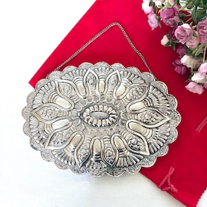 May include: An oval silver-toned metal clutch bag featuring an intricate embossed floral and petal pattern with a scalloped edge. The accessory includes a thin silver-toned metal chain strap for carrying and a central oval textured detail.