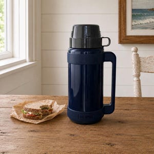 Peut inclure: Une bouteille isotherme de marque Thermos bleu marine foncé avec un couvercle à vis noir servant de tasse, une grande poignée latérale et une base assortie, placée à côté d'un sandwich sur un morceau de papier sulfurisé.