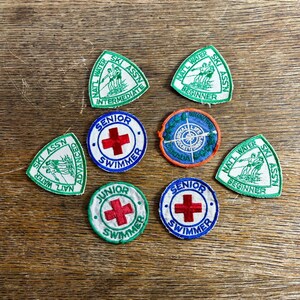 Pode incluir: Uma coleção de patches vintage, incluindo distintivos triangulares verdes e brancos da associação de esqui e distintivos redondos com cruzes vermelhas e as palavras "Senior Swimmer" e "Junior Swimmer". Os patches estão em uma superfície de madeira.