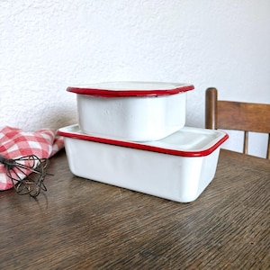 May include: Two rectangular white enamel storage containers with lids, each featuring a thin red rim. The containers are stacked, with the smaller one placed on top of the larger one. The finish is smooth and glossy, showing a classic kitchen design.