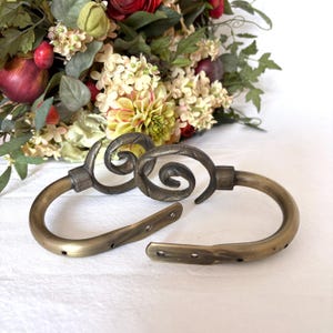 Peut inclure: Deux embrasses de rideaux en laiton antique présentant un design incurvé en demi-cercle avec une volute décorative en spirale sur le dessus. Chaque pièce en métal comprend des extrémités de fixation plates avec des trous pré-percés.