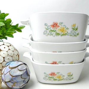 May include: Set of four white ceramic baking dishes with floral designs. The dishes are stacked on top of each other. The floral designs feature pink, yellow, and blue flowers.