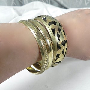 Peut inclure: Ensemble de quatre bracelets en métal doré. Les bracelets sont empilés sur le poignet d'une personne. Le bracelet du haut présente un motif floral découpé.