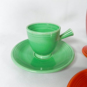 May include: A green ceramic teacup and saucer set. The cup has a handle and a rounded base. The saucer has a scalloped edge.