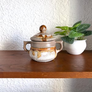 May include: A brown and white ceramic sugar bowl with a lid. The bowl has a drip glaze and a rounded shape. The lid has a knob on top.