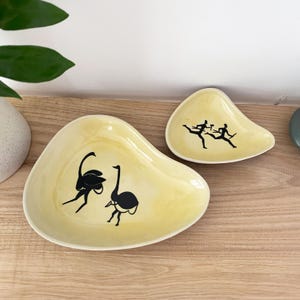 Op de afbeelding: Twee gele, boemerangvormige keramische schalen. De grotere schaal heeft een zwarte illustratie van twee struisvogels, terwijl de kleinere schaal een zwarte illustratie heeft van twee rennende figuren. De schalen staan op een houten oppervlak.