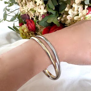 Op de afbeelding: Twee zilveren armbanden, waarvan één goudkleurig, worden om een pols gedragen. De armbanden zijn glad en cirkelvormig. Op de achtergrond een boeket bloemen met rode, witte en groene tinten.