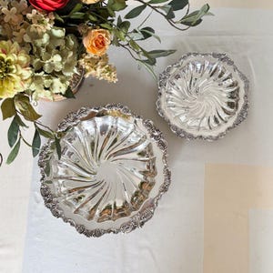May include: Two ornate, silver-colored serving dishes with a spiral design in the center. The dishes have a decorative, scalloped edge. A floral arrangement with orange and yellow flowers is in the background.