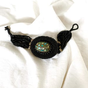 May include: Black beaded necklace with an oval turquoise and stone pendant. The pendant is surrounded by a black beaded border. The necklace has a beaded tassel on each side and a black cord closure.