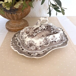 May include: A rectangular ceramic butter dish with a lid featuring a brown and white transferware pattern of floral motifs and architectural elements. The dish has a scalloped edge and a decorative handle on top of the lid with a matching pattern.