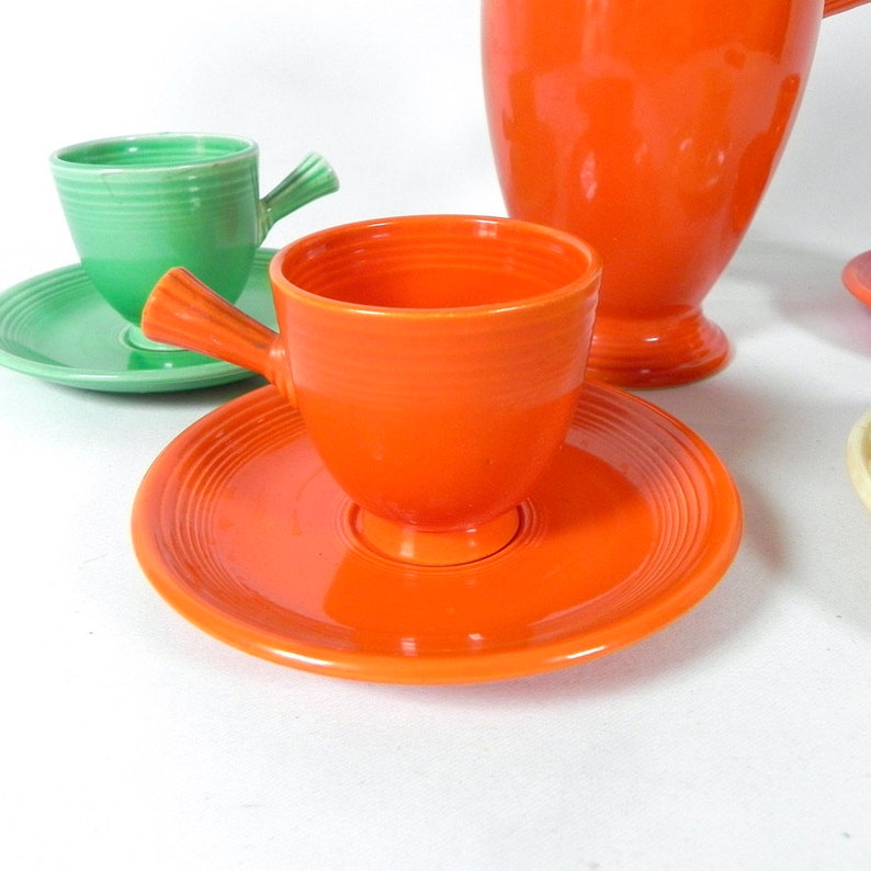 May include: A set of vintage Fiestaware teacups and saucers in orange and green. The cups have a unique handle design and the saucers have a ridged pattern.