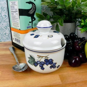 Op de afbeelding: Witte suikerpot van keramiek met een blauwe en gouden rand. De pot heeft een deksel met een klein tuitje en een ontwerp met zwarte bessen. De pot staat op een houten oppervlak.