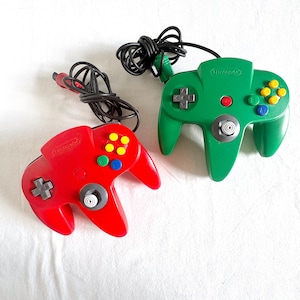 May include: Two Nintendo 64 video game controllers, one in solid red and one in solid green. Each controller features a central analogue stick, a directional pad, four yellow C-buttons, A and B buttons, and a black wired cord with a connector plug.