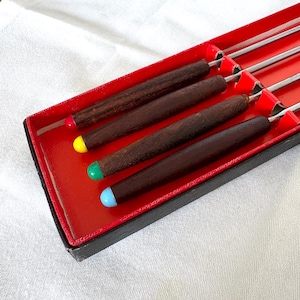 Può includere: Un set di forchette da fonduta in una scatola rossa. Le forchette hanno manici in legno marrone scuro con punte colorate: rosso, giallo, verde e blu. La scatola ha un bordo nero e un interno rosso.