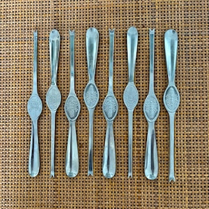 May include: Eight silver fruit forks are displayed on a woven surface. Each fork has an oval handle with an embossed design and a slender tine. The forks are likely vintage or antique, and are made of silver-toned metal.