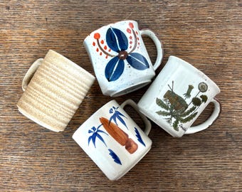 May include: Four ceramic mugs with unique designs. One mug has horizontal tan stripes, while others feature floral and abstract patterns in blue, brown, and green on a white background. Each mug has a brown handle.