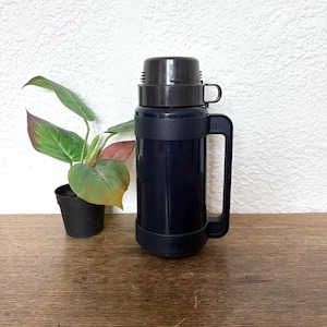 May include: A navy blue Thermos brand insulated bottle with a black lid and handle. The bottle has a black band around the middle and base. A small potted plant with green and red leaves sits to the left of the bottle.
