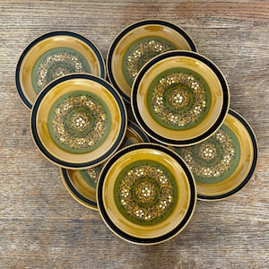 Puede incluir: Un juego de siete platos redondos de cerámica con un círculo central verde oliva rodeado por un borde amarillo mostaza y un borde exterior negro. Cada plato muestra un patrón decorativo de enredaderas negras y pequeños detalles florales blancos.