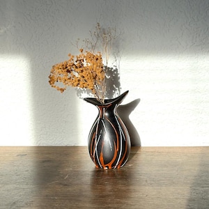 May include: A black ceramic vase with a unique shape and a decorative pattern of orange and white lines. The vase holds dried, tan-colored flowers. The vase is on a wooden surface, with a white wall in the background.