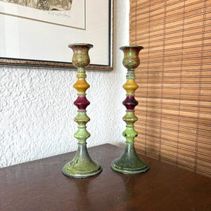 Peut inclure: Deux bougeoirs décoratifs avec une finition tachetée. Les bougeoirs ont une base verte, un centre rouge et une section médiane jaune. Le dessus est de couleur rouge foncé. Les bougeoirs sont sur une surface marron foncé.