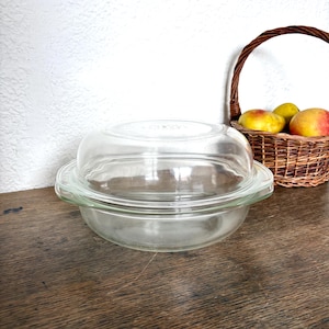 Könnte beinhalten: Eine klare Pyrex-Backform aus Glas mit passendem Deckel. Die Form ist rund mit einem leicht ausgestellten Rand. Ein Weidenkorb mit Äpfeln steht im Hintergrund auf einer Holzoberfläche.