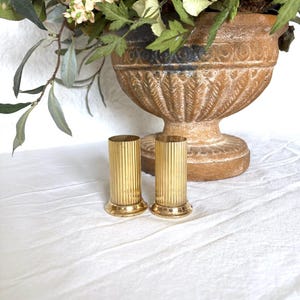 May include: Two gold-colored, fluted candle holders with a round base. The candle holders are cylindrical and stand upright on a white surface. A large, decorative urn with flowers is in the background.