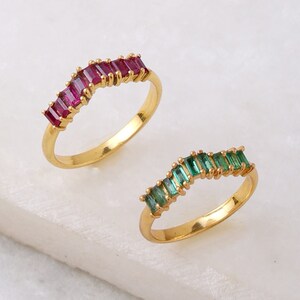May include: Two gold rings with a row of rectangular gemstones. One ring has green gemstones and the other has red gemstones.
