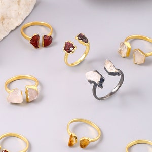May include: Assortment of gold rings with different colored gemstones. The rings feature two stones on each side of the band. The stones are raw and have a natural, rough texture. The rings are a mix of red, orange, and white gemstones.
