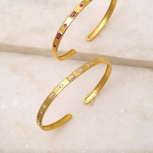 May include: Two gold bangle bracelets with a row of small rectangular gemstones. One bracelet has pink gemstones and the other has clear gemstones.