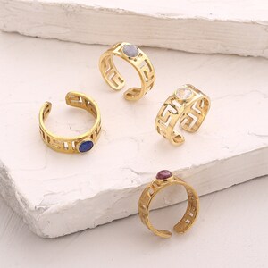 May include: Four gold rings with a geometric design and different colored gemstones. The rings are on a white surface.