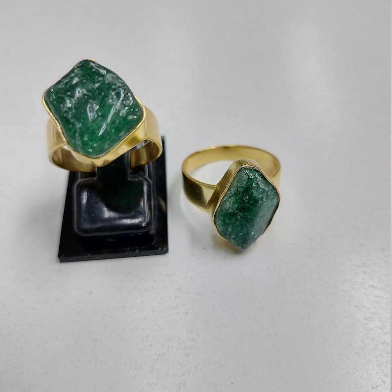 May include: Two gold rings with green gemstone settings. The rings are on a black display stand. The larger ring has a more irregular shaped stone.
