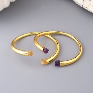 May include: Two gold bangle bracelets with a purple and orange gemstone accent at the end of each bracelet.