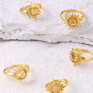 May include: Five gold-colored rings with a sunburst design. Each ring features a light-colored, square-cut gemstone at the center. The rings are displayed on a textured, off-white surface.