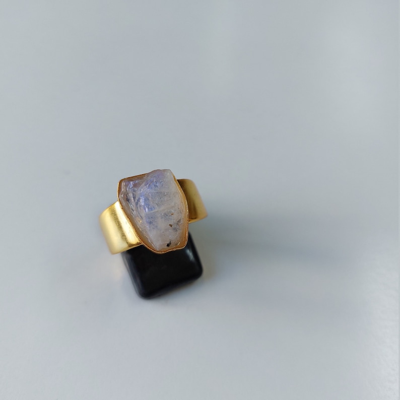 May include: A gold ring with a large, clear, white moonstone set in the center. The ring is on a black display stand.