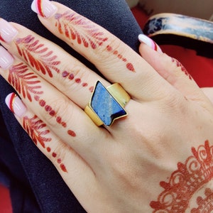 May include: A gold ring with a large, triangular blue gemstone. The ring is on a hand with henna designs.