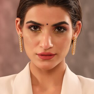May include: A woman wearing a white blazer with gold earrings that have clear stones. The earrings are long and rectangular.
